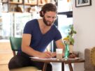 Smiling young man sitting in a coffee shop writing letter
